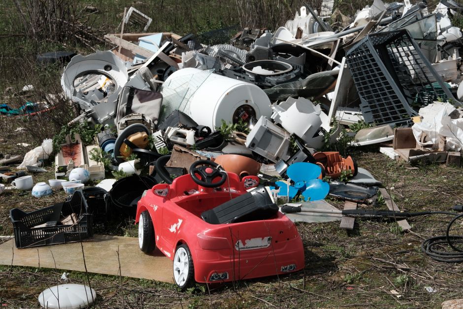 The image depicts a large pile of various discarded household items and debris outdoors, situated on a patch of uneven ground with sparse grass and small plants. Prominently in the foreground, there is a red plastic children's ride-on toy car with a slightly worn appearance, showing some scratches and paint chips on its surface. Surrounding the toy car are scattered objects including a black plastic basket, white ceramic bowls, a blue balloon partially deflated, and a black garden hose lying on the ground. Behind the toy, the bulk of the pile consists of an assortment of household appliance parts, including a white washing machine drum, metallic and plastic casing pieces, and damaged kitchenware. These items display various textures, from shiny and smooth plastics to matte and weathered metals, with some components showing evidence of dirt and corrosion. The background shows a mix of dry brush and trees, indicating a rural or semi-rural environment, and the scene appears to be a site for waste collection or site clearance, related to private rubbish disposal services. The natural lighting highlights the different materials and the disorderly arrangement of discarded objects typical of non-regulated waste dumping areas.