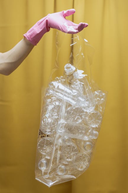 A person's arm, wearing a pink rubber glove, is visible holding a large, transparent plastic bag filled with crushed plastic bottles and containers. The plastic items inside the bag are clear, with some showing ridged or smooth surfaces, and are of varying sizes and shapes. The background features a plain, yellow curtain or wall, providing a neutral backdrop that emphasizes the plastic waste being held. The scene appears to relate to waste collection or recycling, aligning with the services offered by Rubbish Clearance Ilford, particularly in the context of alternative waste handling or on-site rubbish removal. The lighting is even, highlighting the clarity and texture of the plastic debris. Overall, the image captures a moment of managing plastic waste, which is relevant to private rubbish disposal services that focus on environmentally responsible clearance and recycling practices.