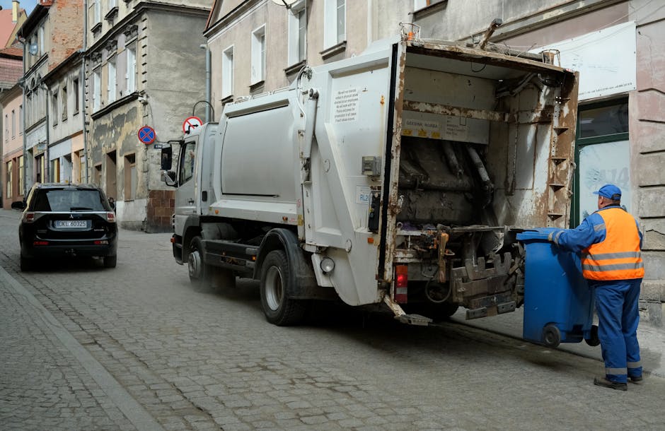 A street scene showing a white commercial rubbish collection truck parked alongside a row of older, weathered buildings with textured stone and concrete facades. The truck's rear compaction section is open, revealing rusted metal surfaces and mechanical components used for compressing waste. A worker dressed in a high-visibility orange vest, blue uniform, and blue cap is positioned on the right side of the image, holding a blue wheelie bin and preparing to dispose of waste into the truck. To the left, a black car is parked on the cobblestone street, which is bordered by a narrow pavement. In the background, several no parking signs are affixed to the façades, and natural daylight illuminates the scene, emphasizing the urban environment associated with independent waste disposal and rubbish removal services provided by Rubbish Clearance Ilford.
