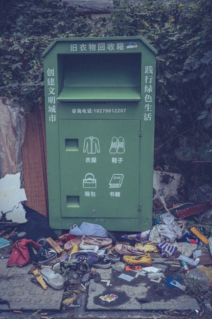 A green clothing donation and recycling bank positioned outdoors against a backdrop of dense foliage and a wooden fence visible on the left side. The bank features a large rectangular opening at the top for depositing used clothing, along with a contact telephone number printed on the front. Below this, there are four smaller compartments labeled with Chinese characters, indicating locations for clothing, shoes, bags, and books. Surrounding the base of the bank is a significant accumulation of discarded items, including various shoes, clothing, paper, plastic wrappers, and other miscellaneous rubbish scattered across the ground. The environment appears untidy, with trash spilling out from the bins and laying on the pavement, contributing to visual clutter. The scene reflects a situation where a designated waste or donation container is overwhelmed or improperly used, highlighting challenges connected to private rubbish clearance and disposal. The image subtly underscores the importance of proper waste management and the role of waste removal services like Rubbish Clearance Ilford in maintaining cleanliness and supporting sustainable disposal practices.