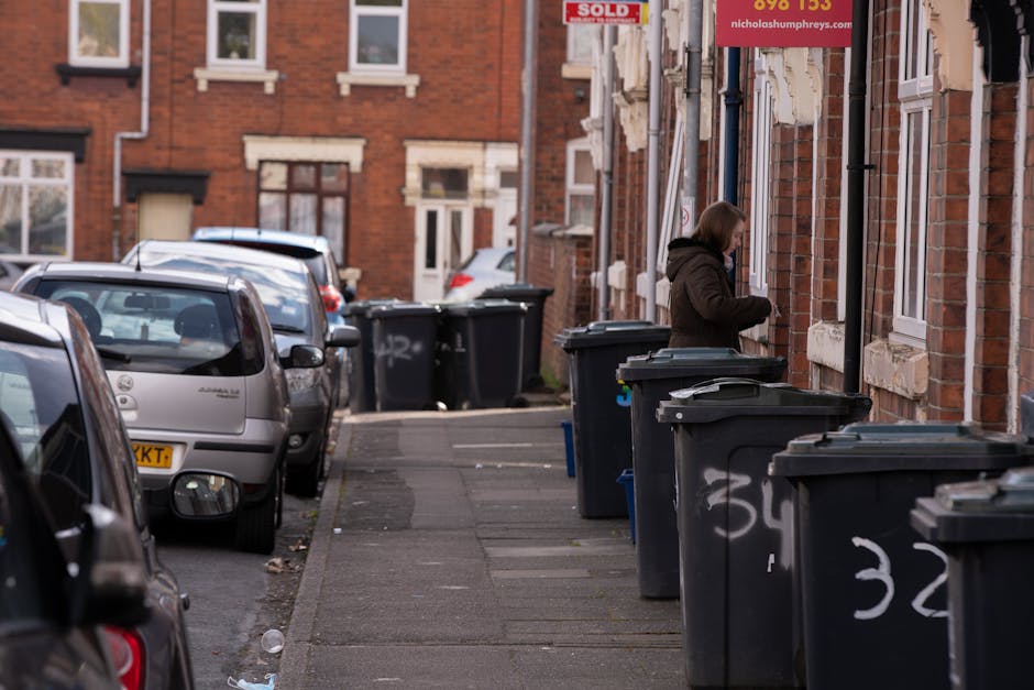 A woman standing on a narrow sidewalk next to a row of large, dark grey wheelie bins with white painted numbers, positioned in front of a red brick residential building. The bins are lined up along the building’s exterior wall, which features multiple white-framed windows and white decorative stone sills. The woman appears to be opening or closing the lid of one of the bins, dressed in a dark brown jacket with a fur-lined hood. Several parked cars are visible on the street to the left, including a silver hatchback closest to the foreground. The street is paved with asphalt, and the sidewalk is made of concrete slabs, some of which have small stains or patches. In the background, additional bins and part of a street with similar houses can be seen, with a red and white sign mounted on the building indicating property sale. The lighting suggests it is daytime with overcast or diffused natural light, and the scene reflects an everyday street scene involving private refuse storage and collection in an urban area, consistent with non-local authority waste management practices.