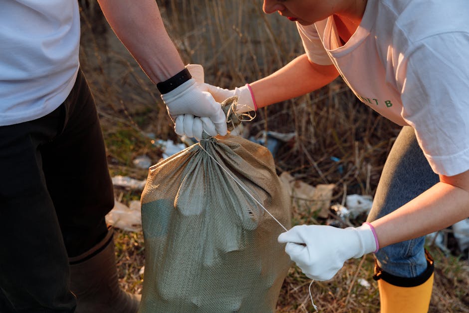 A close-up view of two individuals wearing white gloves, working outdoors during daylight hours. One person, wearing a short-sleeved light-colored shirt and dark pants, holds a large, light brown, woven plastic sack filled with various waste items. The other individual, dressed in a white T-shirt and blue jeans, is gripping a wire or twine attached to the sack, possibly securing or closing it for rubbish removal. The background features a natural environment with dry, brown vegetation and scattered litter, including plastic and miscellaneous debris, indicating a cleanup or waste collection effort in an overgrown, possibly neglected outdoor area. The scene exemplifies on-site waste handling and the importance of responsible rubbish disposal, aligning with services like those offered by Rubbish Clearance Ilford, which focus on independent collection and private waste clearance during rubbish removal tasks.