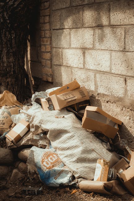 A pile of assorted waste materials, including flattened cardboard boxes, paper bags, and a large white sack, is situated on the ground next to a brick wall and a wooden tree trunk. The cardboard boxes vary in size and are primarily light brown, some with visible labels and printed barcodes. The white sack appears to be woven and is partially filled, with some waste spilling over the top. The ground beneath the waste is uneven, composed of dirt and small rocks. Behind the pile, the brick wall is made of concrete blocks with a textured, rough surface, and the bricks are arranged in a horizontal pattern with visible mortar joints. To the left, a large tree trunk with dark, textured bark extends upwards, partially shaded. The scene is naturally lit, casting soft shadows and highlighting the textures of the waste and surrounding environment. This image reflects concepts of private waste disposal and rubbish removal, as managed by professional services such as Rubbish Clearance Ilford, and depicts an example of on-site clearance often handled outside of municipal collection.