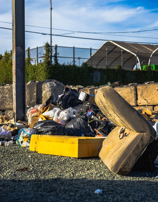 An outdoor scene showing a pile of mixed waste consisting of black and transparent plastic rubbish bags, some partially torn open revealing contents such as paper, packaging, and small debris, alongside an old, worn car tyre lying on its side. The waste is situated on a gravel surface adjacent to a low stone wall constructed of roughly squared stones. In the background, there is a tall metal pole with wiring attached, and behind it, a dense hedge with green foliage. Further behind, a large covered sports court with a curved roof and metal supports is visible, partially obscured by the hedge. The lighting suggests daytime with clear, partly cloudy sky, emphasizing the contrast between the waste materials and the surrounding environment. This scene illustrates a typical scenario where private waste collection and rubbish removal services, such as those provided by Rubbish Clearance Ilford, are often needed to manage clutter like this in residential or urban settings, especially when dealing with non-standard, on-site waste disposal methods.
