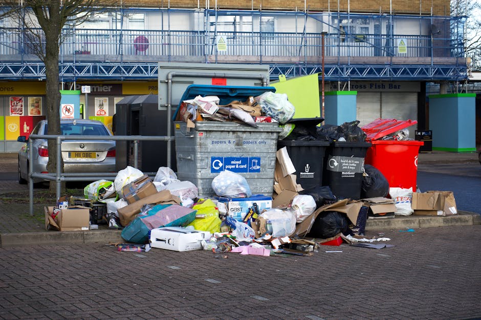 A pile of assorted waste materials, including flattened cardboard boxes, paper bags, and a large white sack, is situated on the ground next to a brick wall and a wooden tree trunk. The cardboard boxes vary in size and are primarily light brown, some with visible labels and printed barcodes. The white sack appears to be woven and is partially filled, with some waste spilling over the top. The ground beneath the waste is uneven, composed of dirt and small rocks. Behind the pile, the brick wall is made of concrete blocks with a textured, rough surface, and the bricks are arranged in a horizontal pattern with visible mortar joints. To the left, a large tree trunk with dark, textured bark extends upwards, partially shaded. The scene is naturally lit, casting soft shadows and highlighting the textures of the waste and surrounding environment. This image reflects concepts of private waste disposal and rubbish removal, as managed by professional services such as Rubbish Clearance Ilford, and depicts an example of on-site clearance often handled outside of municipal collection.