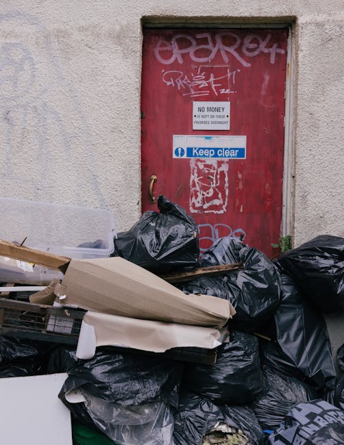 A closed red metal door with graffiti and signs, including a white sign reading 'NO MONEY' and a blue sticker that says 'Keep clear,' is set into a rough-textured, beige concrete wall. In front of the door, there is a large pile of black rubbish bags, some torn open, with materials such as cardboard, plastic, and paper partially visible among the accumulative waste. The rubbish is positioned on a paved surface, which appears to be a narrow alley or driveway, suggesting a private or back-entry area for waste collection or disposal. The scene is illuminated by natural light, giving the area a neutral, unkept appearance typical of informal rubbish accumulation. The presence of the rubbish bags and discarded materials highlights a context of waste management or rubbish clearance, which fits with services offered by companies like Rubbish Clearance Ilford for alternative waste handling solutions outside regular municipal collection.