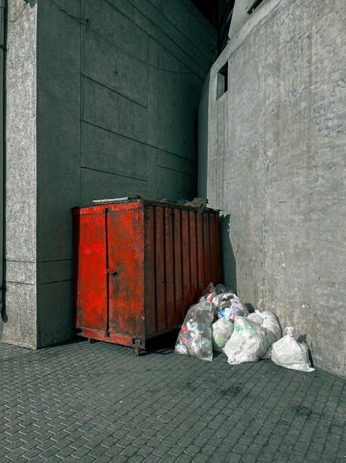 A white ceramic toilet bowl, which appears to be disused and discarded, is situated outdoors beside a blue metal gate and a dark brown fence. The toilet is surrounded by a variety of litter, including crumpled paper, plastic bottles, takeaway containers, and general rubbish cluttering the ground around it. Several empty bottles, some plastic, are stacked or scattered on and around the toilet, with one green glass bottle standing upright on the rim. A partially crushed paper bag and other miscellaneous debris are also visible among the trash. The concrete surface beneath the litter shows signs of dirt and stains, with some cracks and wear. The scene suggests an environment in need of private waste removal or on-site clearance, with the clutter indicative of informal or unmanaged rubbish disposal practices, which Rubbish Clearance Ilford could assist in managing.