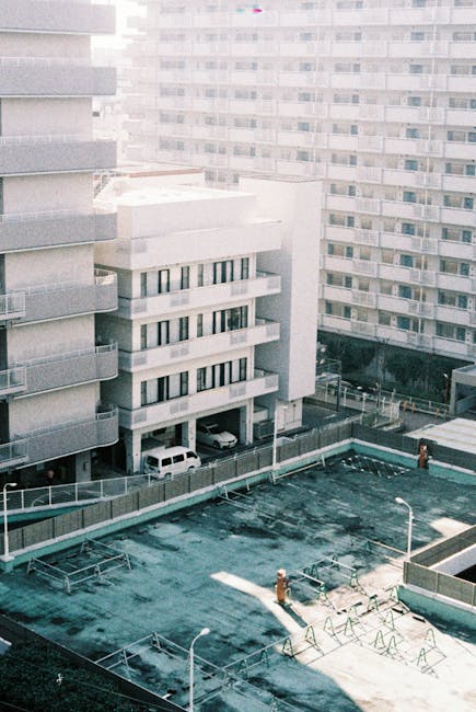 An aerial view of a multi-storey residential building with a light grey facade, featuring several balconies with horizontal railings and numerous windows, situated adjacent to other similar high-rise blocks. In the foreground, there is a flat rooftop area with a worn, dark green surface that appears to have some patches or discoloration, surrounded by a railing and equipped with a few small, upright structures, possibly for maintenance or safety. Below the building, a parking area accommodates a few parked vehicles, including a white van and a white car, parked directly under the building's overhang. The overall setting reflects an urban environment with modern apartment complexes. The scene, captured in natural daylight, emphasizes the building's architecture and the rooftop's utilitarian appearance, subtly contextualizing the type of environment where efficient rubbish removal services by companies like Rubbish Clearance Ilford are often needed for managing waste from high-density residential areas close to commercial or private disposal options.