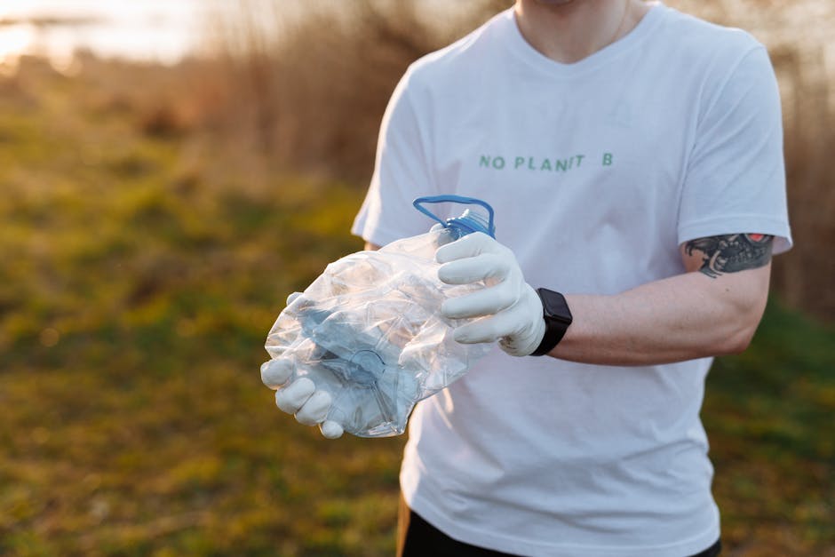 A person wearing a white T-shirt with the text 'NO PLANT B' printed in green, dressed in white gloves, is holding a clear plastic bag filled with crumpled white and beige materials, possibly paper or fabric scraps. The individual is outdoors in a natural setting, with a blurred background of grass and shrubbery illuminated by soft, warm light, suggesting late afternoon or early evening. The person’s left arm, which is visible, features a black wristwatch and a partially visible tattoo on the forearm. The scene appears to relate to waste collection or environmental activities, with the plastic bag potentially prepared for disposal or recycling in the context of private waste handling or rubbish removal services. The focus is on the hands and the bag, highlighting careful handling of waste materials, consistent with professional rubbish clearance practices carried out by companies such as Rubbish Clearance Ilford.