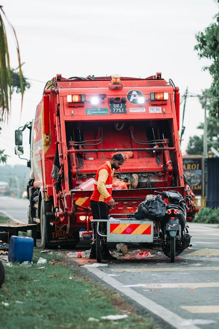 A worker in a red and yellow uniform operates a large red rubbish collection truck parked on the side of a street during daytime. The rear loading mechanism of the truck is open, revealing internal components and the hydraulic system. The worker is handling waste items, including a black garbage bag, which are positioned near the truck's rear platform. The street setting includes a paved sidewalk, green grassy areas, and nearby street furniture such as a black motorcycle parked close to the truck. In the background, there are trees and a building with signage, indicating a semi-urban environment. The sky appears overcast, providing diffuse natural lighting. This scene illustrates an example of private waste disposal or alternative rubbish removal services, aligning with the topic of on-site cleanup and independent collection methods facilitated by companies like Rubbish Clearance Ilford.