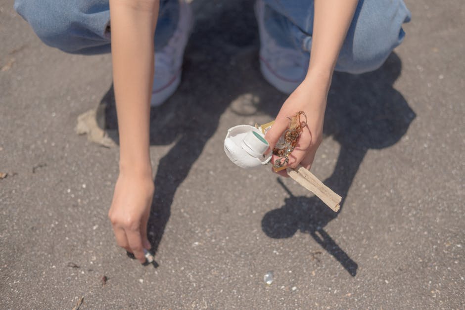 A person wearing a white T-shirt with the text 'NO PLANT B' printed in green, dressed in white gloves, is holding a clear plastic bag filled with crumpled white and beige materials, possibly paper or fabric scraps. The individual is outdoors in a natural setting, with a blurred background of grass and shrubbery illuminated by soft, warm light, suggesting late afternoon or early evening. The person’s left arm, which is visible, features a black wristwatch and a partially visible tattoo on the forearm. The scene appears to relate to waste collection or environmental activities, with the plastic bag potentially prepared for disposal or recycling in the context of private waste handling or rubbish removal services. The focus is on the hands and the bag, highlighting careful handling of waste materials, consistent with professional rubbish clearance practices carried out by companies such as Rubbish Clearance Ilford.